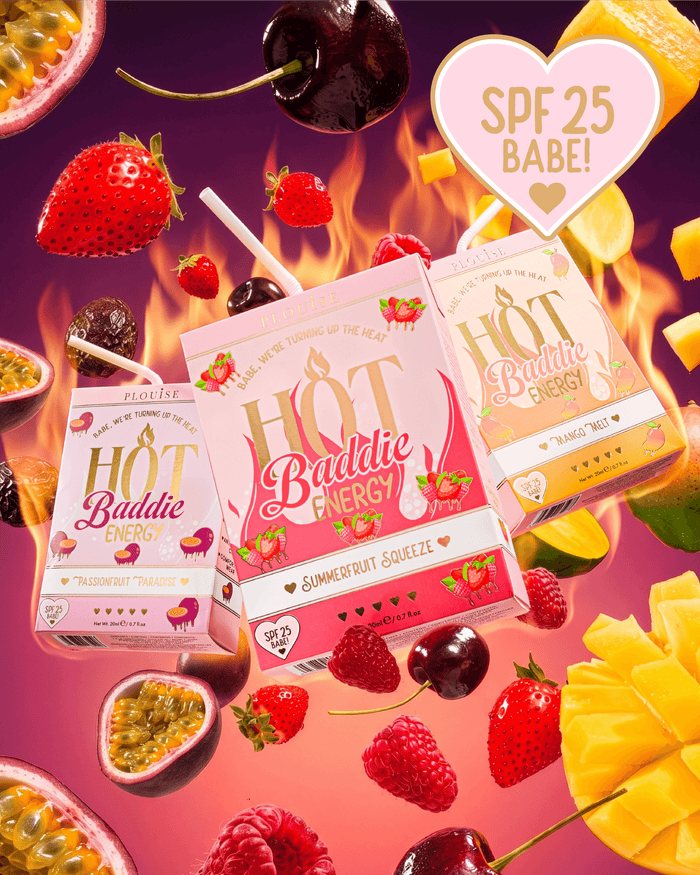 Three juice boxes from Hot Baddie Energy SPF Lip Conditioner and Lip Oil Duo are arranged in a dynamic floating composition surrounded by fresh fruit slices, cherries, strawberries, and passion fruit on a vibrant pink and orange background with flame graphics. 
