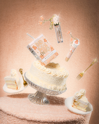 टेडी फैब्रिक टेबल पर एक केक, जिसमें नारियल के केक में पी.लुईस टेडी ग्लिटर बैड बिच एनर्जी लिप डुओ है, जिसमें लिप कंडीशनर और केक के चारों ओर लिप ऑयल तैर रहा है। 